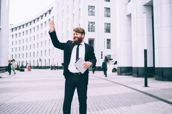 Friendly best man in elegant suit with tablet in hands waving hand while standing near magnificent building of banquet hall