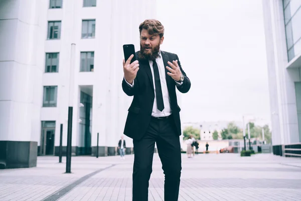 Bearded man with brown hair in black suit and tie standing on street having video call via smartphone and gesticulating