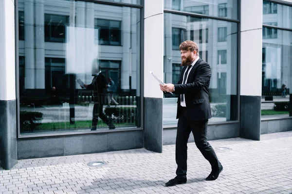現代的なオフィスの近くの舗装に沿って歩いている間エレガントな服やネクタイチェック文書で完全な長さの高価なひげそりのビジネスマンの側面図 — ストック写真