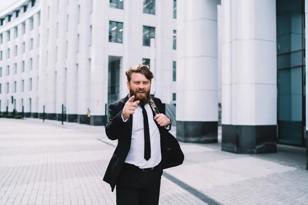 Cheerful young man in formal wear with beard pointing with finger and looking at camera with bright smile while walking along street