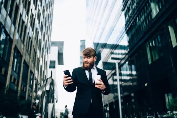 Focused bearded entrepreneur with cup of coffee standing on light city street near contemporary glass buildings and browsing internet on smartphone