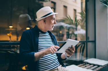 Güncel kafenin penceresinin yanında oturan ve tableti incelerken yüzünü çeviren, sıradan kıyafetli ve şapkalı dalgın bir son sınıf öğrencisi.