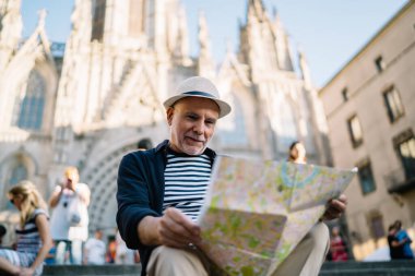 Gülümseyen, modaya uygun şapkalı yaşlı bir adamın haritaya bakıp antik bir katedralin yakınındaki merdivenlerde oturmasından.