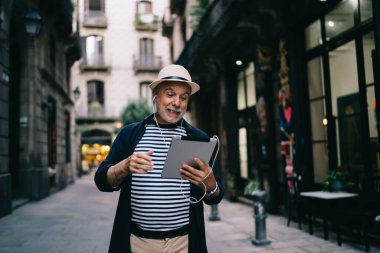 Günlük giysiler içinde gülümseyen yaşlı bir erkek. Modern şehir caddesinde yürüyorlar. Tabletten video aracılığıyla konuşuyorlar.