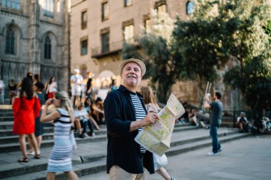 Normal giyinmiş, şapkalı, gülümseyen olgun erkek turistin eski turistik yerlerin yakınındaki caddenin ortasında elinde haritayla duruşu.