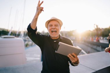 50 yaşındaki neşeli erkek elinde tabletle şehrin deniz kenarında dururken mutlu bir şekilde gülümsüyor.