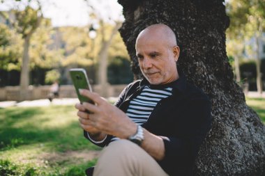 Sıradan kıyafetlerle kel kafalı, odaklanmış son sınıf öğrencisi yere oturmuş ve cep telefonuna bakarken ağaca yaslanmış.