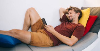 Hafta sonu modern akıllı telefonuyla rahat bir kanepede rahat rahat uyuyan mutlu genç adamın yan görüntüsü.