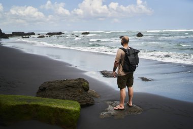 Yüksek açılı, kimliği belirsiz, sıradan kıyafetleri ve sırt çantasıyla gezen, fırtınalı deniz manzarasının keyfini çıkaran erkek yolcu.