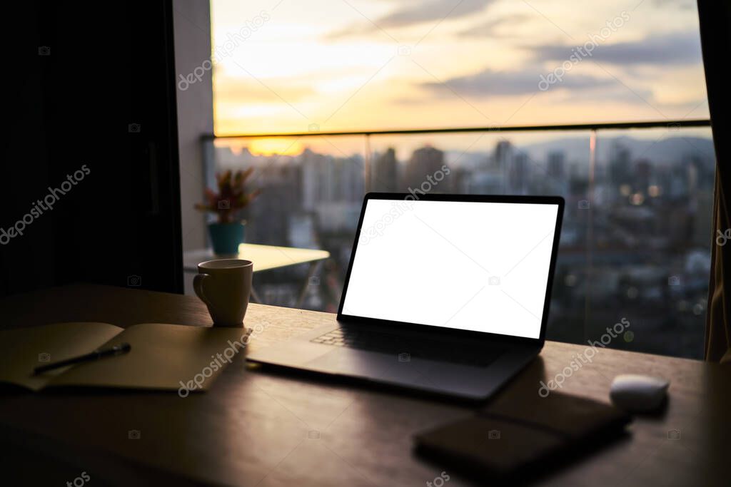 Contemporary workspace with blank screen netbook and planner placed on wooden table near window of skyscraper overlooking downtown during sunset