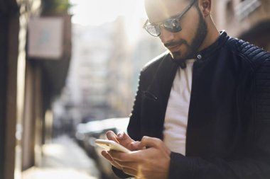 Moda kıyafetleri ve güneş gözlüklü sakallı bir erkek gündüz sokakta dururken sosyal medyada cep telefonuyla geziniyor.