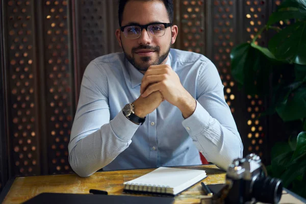 Confident male in formal wear and eyeglasses looking at camera while sitting at desk near plant with notepad and camera