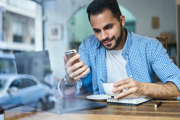 カフェやブラウジングスマートフォンに座ってホットコーヒーのカップとカジュアルな服の正の髭の男性のウィンドウを介して — ストック写真