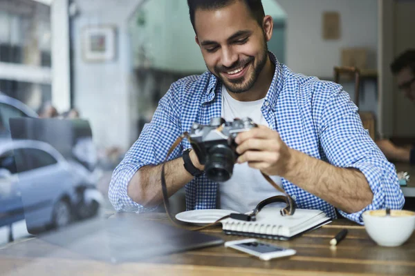 幸せな髭を生やした男性は ノートパソコンで作業しながら コーヒーとコピーブックのカップとテーブルに座って写真カメラでカジュアルな服を着ています — ストック写真