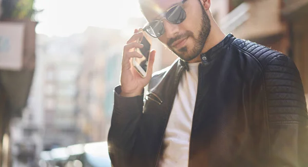 身着时髦皮夹克 戴着太阳镜站在路边 一边在手机上听留言 一边空余时间的作物族男性 — 图库照片