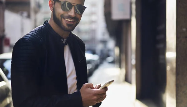 街の通りの近代的な建物を過ぎて歩いている間 スマートフォンでスタイリッシュな衣装メッセージでハンサムな男性をクロップし カメラで微笑む — ストック写真