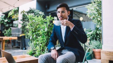 Gözlüklü, resmi takım elbiseli, kahve içen ve modern sokak kafesinde dinlenirken kameraya bakan neşeli Asyalı adam.