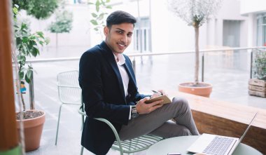 Şık takım elbiseli, kendine güveni tam bir etnik adamın masada bacak bacak bacak üstüne atmış bir şekilde oturması ve modern sokak kafesinde öğle yemeği arasında dizüstü bilgisayarla çalışırken not alması.