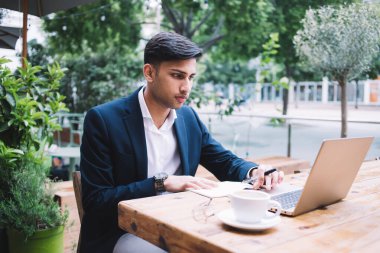 Zarif giyinen konsantre etnik genç adam ekrana bakarak ve not defterine not alarak dışarıda kafede bir fincan kahveyle çalışırken