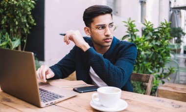 Gözlüklü genç etnik bir adam, laptopla ve kafeterya terasında bir fincan kahveyle çalışırken dikkatle başka tarafa bakıyor.
