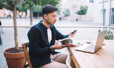 Kaldırımdaki kafede dizüstü bilgisayarla çalışırken akıllı telefon kullanarak kitap çeken genç bir adamın yan görüntüsü