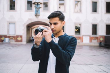 Tarihi binanın bulanık arka planında dikilirken dijital kamerada fotoğraf çeken şık giyinmiş genç etnik erkek fotoğrafçı.