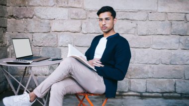 Moda kıyafetli ciddi bir etnik adam kafeteryada dizüstü bilgisayar çalışırken bacak bacak bacak üstüne atmış bir şekilde oturuyor ve dergi okuyor.