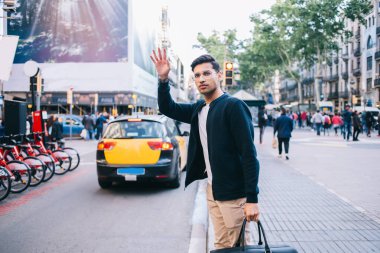Güneşli bir günde kaldırımda dikilirken taksiye binmek için elini kaldıran gözlüklü ciddi bir adam.