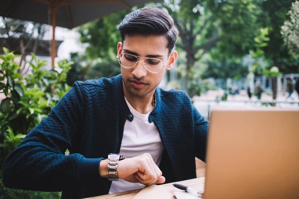 穿着时髦眼镜的严重落后的男性创业者 一边在咖啡店的阳台上工作一边检查视力 一边坐着沉思沉思 拿着笔记本电脑看手表 — 图库照片