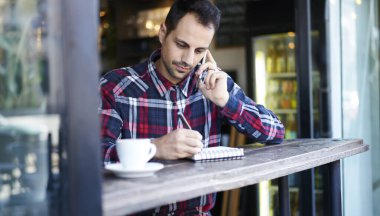Düşünceli, günlük kıyafetlerle serbest çalışan bir erkek, masada oturuyor ve kafeteryada akıllı telefonla konuşurken defterine not alıyor.