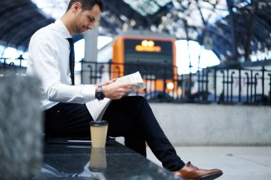 Resmi kıyafetli ciddi bir erkek işçinin, elinde gazeteyle bankta oturup tren istasyonunu beklemesinin yan görüntüsü.