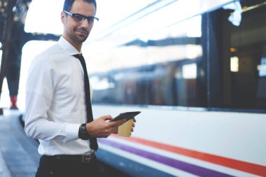 Resmi kıyafetler ve gözlüklerle gülümseyen yetişkin ofis çalışanı tren istasyonunda beklerken akıllı telefon kullanıyor ve kahve içiyor.