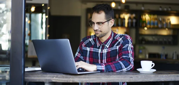 穿着休闲装 头戴眼镜 头脑清醒的男性自由职业者坐在咖啡店的桌子旁 在笔记本电脑上做远程项目 — 图库照片