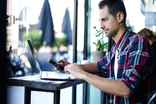身着休闲装 头戴手提电脑坐在木制桌旁 一边在咖啡店用智能手机聊天的男性侧视图 — 图库照片