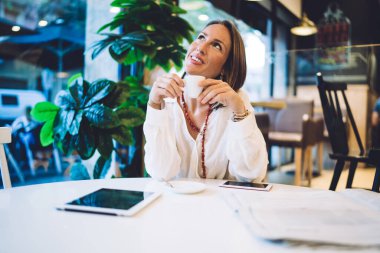 Düşünceli bir kadın kahve molası veriyor. Kafede uzaktan kumandalı cihazlar ve gazetelerle çalışıyor. Düşünceli bir şekilde yukarı bakıyor.