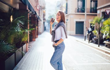 Güneşli bir günde kaldırımda dikilirken günlük kıyafetleriyle gülümseyen ve paket kahve içen pozitif kadın.