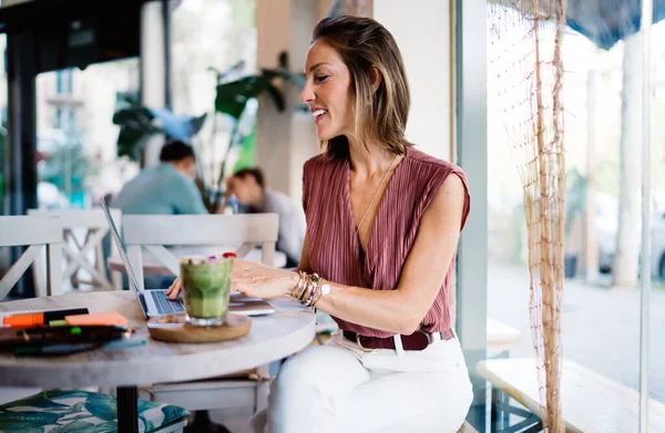 陽気な大人の女性は 朝食のためのスムージーを持っている窓の近くのテーブルに座ってカフェからオンラインで働くネット上で入力 — ストック写真