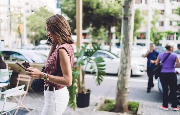 Pretty caucasian businesswoman in trendy wear walking on street in town talking on mobile phone,cheerful female satisfied with tariff and roaming connection strolling on city street making call