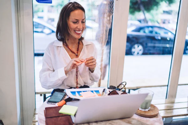 积极的中年女性 穿着雅致的服装 一边用文件和贴纸 一边浏览笔记本电脑 一边在日间舒适的咖啡店里做项目 — 图库照片