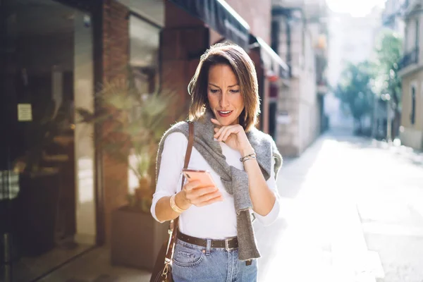 晴れた日には暗い都会の環境に対してスマートフォンでメッセージを読みながら カジュアルな服であごに触れる思考中年齢女性 — ストック写真