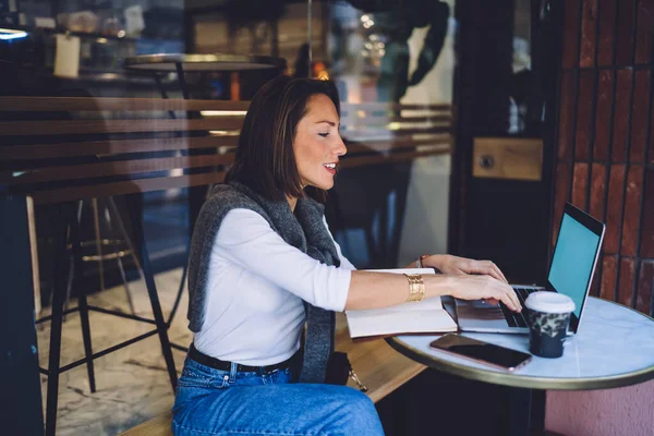 屋外カフェでコーヒーとテーブルに座っている間仕事のためのラップトップを使用してカジュアル服で焦点を当てた中年女性の側面図 — ストック写真
