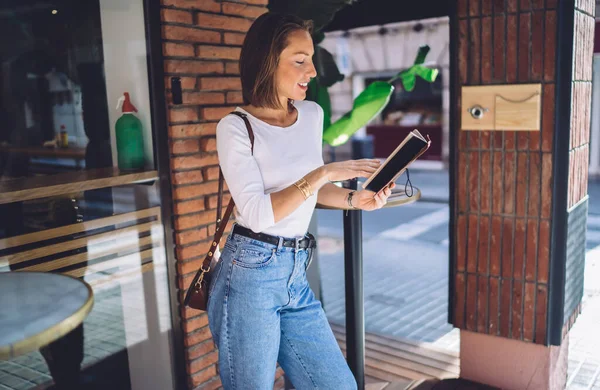 街のカフェの外の通りに立っている間に小さなハンドバッグ読書ノートブックとカジュアルな服装で良い外観の女性の側面図 — ストック写真