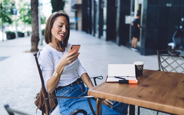 夏のカフェでコーヒーブレイクを楽しみながら 白いシャツと青いジーンズの幸せな女性起業家は 電話でテキストメッセージとブラウジングインターネットを読んで — ストック写真