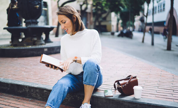 Thoughtful 40 years old woman in trendy outfit reading information in notebook while sitting on stone steps in park during coffee break