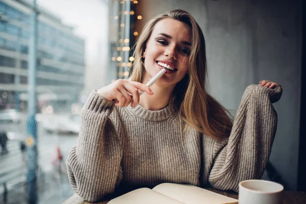 Portrait of happy Caucasian woman with perfect veneers smile looking at camera during learning time, cheerful hipster girl doing school homework in coffee shop sitting at table with education textbook