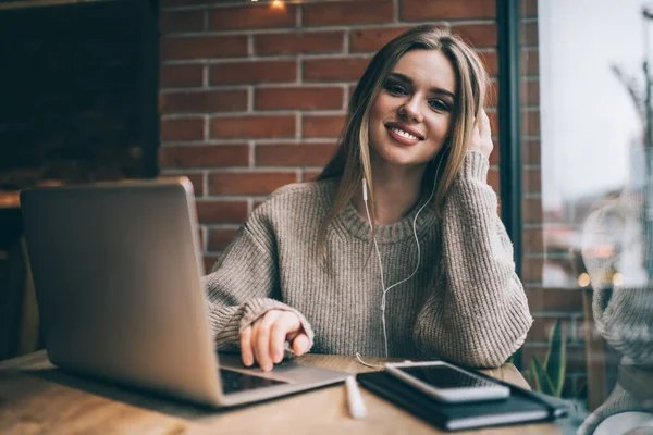 年轻快乐的女性 一头金黄色的长发 穿着针织毛衣 笑着和笔记本电脑一起工作 坐在靠窗的桌旁 看着相机 — 图库照片