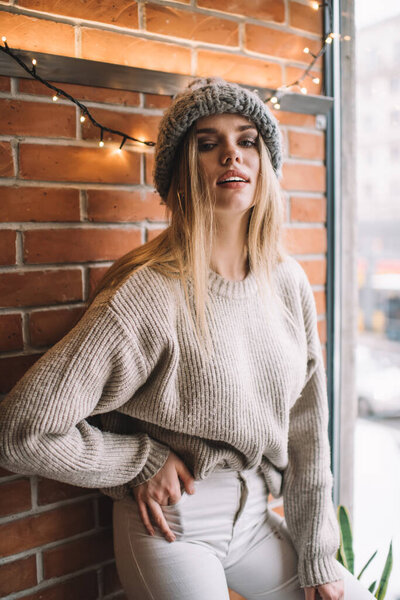 Полудлинный портрет привлекательной белой женщины в трикотажной одежде, стоящей у кирпичной стены и смотрящей в камеру, красивой модели 20 лет в зимней шляпе и пуловере, позирующей в помещении