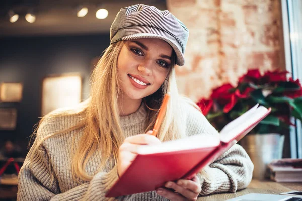 在舒适的咖啡店里 穿着时髦帽子和毛衣的快乐的年轻女士一边在写日记一边看着相机 一边在舒适的咖啡店里休息 — 图库照片