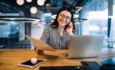 Asyalı, siyah saçlı, gözlüklü, siyah beyaz tişörtlü, dişlek gülüşlü, ofiste otururken telefonla konuşan bir kadın.