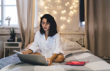 Şık gözlüklü, beyaz gömlekli, dizüstü bilgisayarının üstünde oturmuş, duvarda süslü çelenklerle süslenmiş bir kadın.